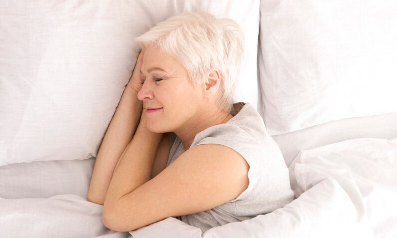 Eine lächelnde Frau mit weißem Haar schläft entspannt auf der Seite in einem weißen Bett. | © Adobe Stock/ Prostock-studio