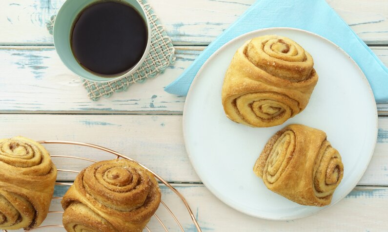 Franzbrötchen ohne Zucker angerichtet auf weißem Teller, daneben weitere Franzbrötchen und eine Tasse Kaffee. | © Getty Images / Westend61