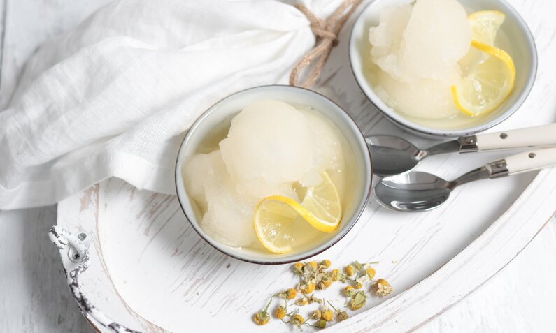 In zwei weißen Schalen angerichtetes Zitronensorbet, daneben frische Zitronenscheiben und frische Minze. | © Getty Images / MelanieMaya