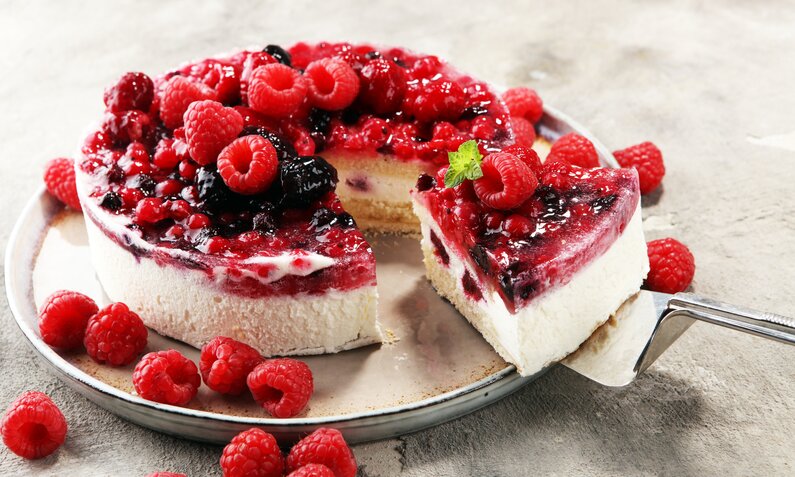 Angeschnittener Joghurt-Himbeerkuchen mit frischen Beeren daneben, angerichtet auf einem Kuchenteller. | © Getty Images / 	beats3