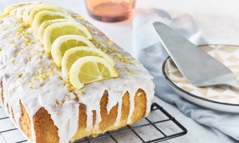 Angeschnittener Zitronenkuchen mit Joghurtguss auf einem Abkühlgitter, daneben Tortenheber auf einem Teller. | © Getty Images / Tim Young Photography