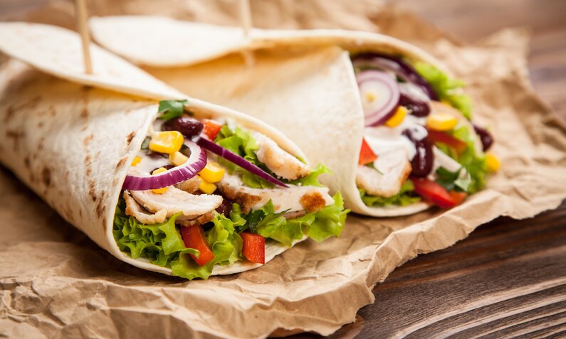 Zwei Wraps mit Hähnchen und Salatfüllung liegen auf Backpapier auf einem Holzuntergrund. | © Adobe Stock / George Dolgikh