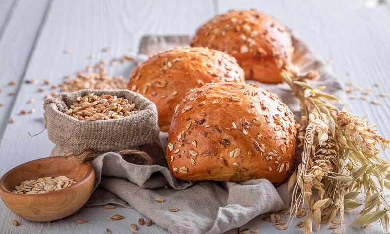 drei Haferbrötchen auf einer Holzunterlage daneben Weizenzweige und Körner. | © Adobe Stock / shaiith