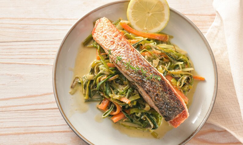 Gebratener Lachs auf Zucchini-Pasta mit Zitronen-Soße auf einem weißen Teller angerichtet. | © Adobe Stock / Maren Winter