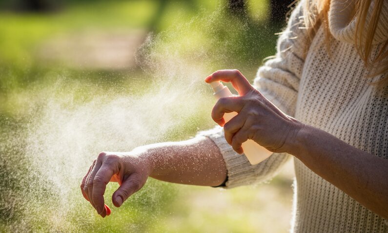 Eine Frau in weißem Strickpullover sprüht sich im Sonnenschein Insektenspray auf den Unterarm. | © Adobe Stock/ encierro