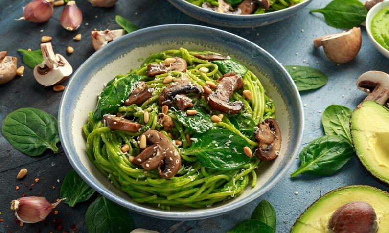 Spinat-pasta auf einem Teller angerichtet, daneben aufgeschnittene Avocado und frischer Babyspinat. | © Adobe Stock / Nick van Bree