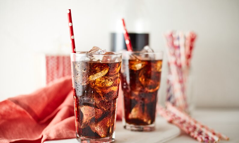 Zwei Gläser Cola mit Eis und rot-weiß gepunkteten Strohhalmen stehen vor einem roten Tuch auf weißem Tisch. | © Adobe Stock/ musicphone1