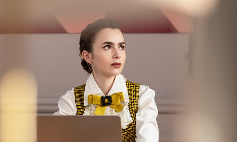 Nahaufnahme von Lily Collins als Emily, die im schicken Outfit in einer Szene aus der Netflix-Serie "Emily in Paris" - Staffel 4 sinnierend hinterm Laptop sitzt. | © Netflix/Marie Etchegoyen