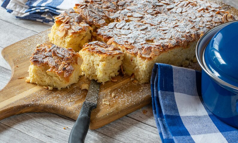 Joghurtkuchen vom Blech auf einem Holzbrett liegend und einem Stück abgeschnitten, on top Mandelsplitter. | © Getty Images / Angelika Heine