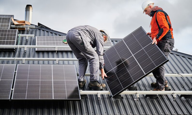 Zwei Handwerker installieren Solarpaneele auf einem schwarzen Metalldach unter bewölktem Himmel. | © Getty Images/anatoliy_gleb