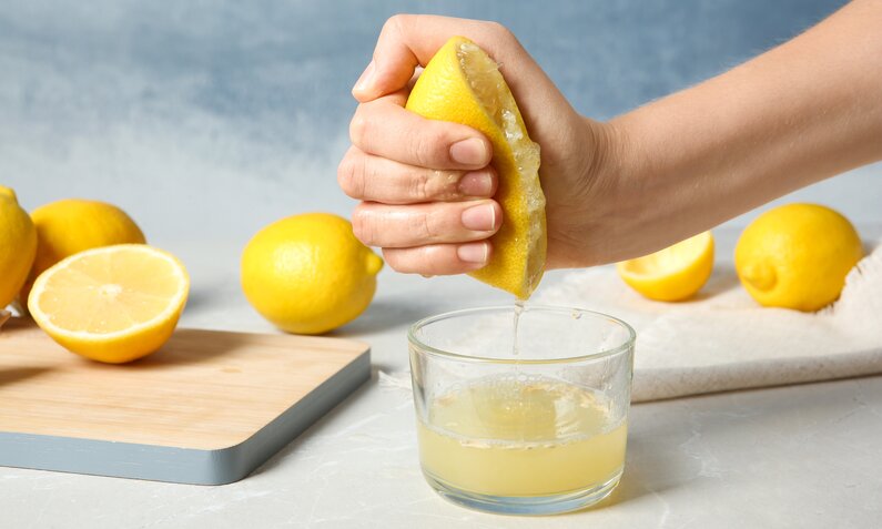 Eine Hand presst eine halbe Zitrone aus, während der Saft in ein Glas tropft, daneben liegen ganze und halbe Zitronen auf einem Schneidebrett. | © Adobe Stock/ New Africa