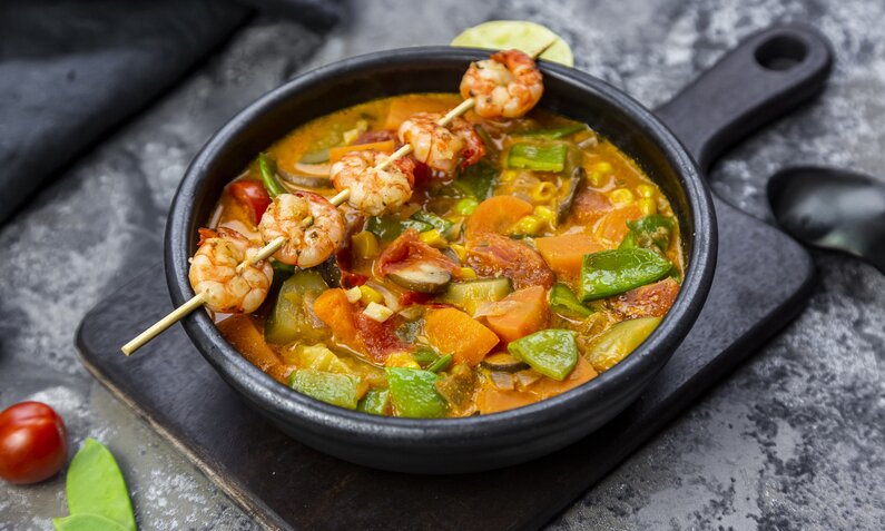 Zucchini-Curry mit Gemüse und Garnelen-Spieß in dunkler Schüssel angerichtet. | © Getty Images / lacaosa