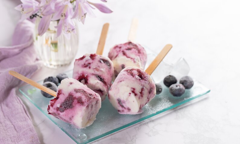 Vier kleine Joghurteis am Stiel mit Blaubeeren, daneben frische Blaubeeren in fliederfarbener Umgebung. | © Getty Images / yumehana