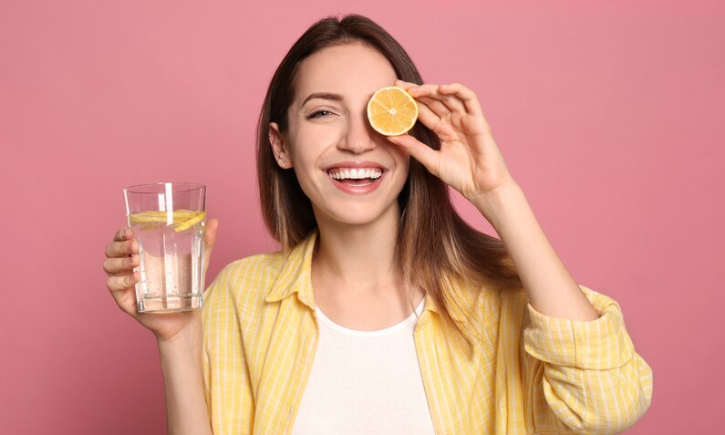 Junge Frau mit braunen Haaren und gelber Bluse hält ein Glas Zitronenwasser und lächelt, während sie eine Zitronenscheibe vor ihr Auge hält. | © Adobe Stock/ New Africa