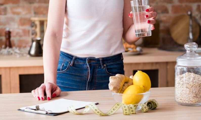 Eine Frau in weißem Top und Jeans hält ein Glas Wasser in der Küche, neben ihr liegen Zitronen, Ingwer und ein Maßband auf dem Tisch. | © Adobe Stock/ golubovy