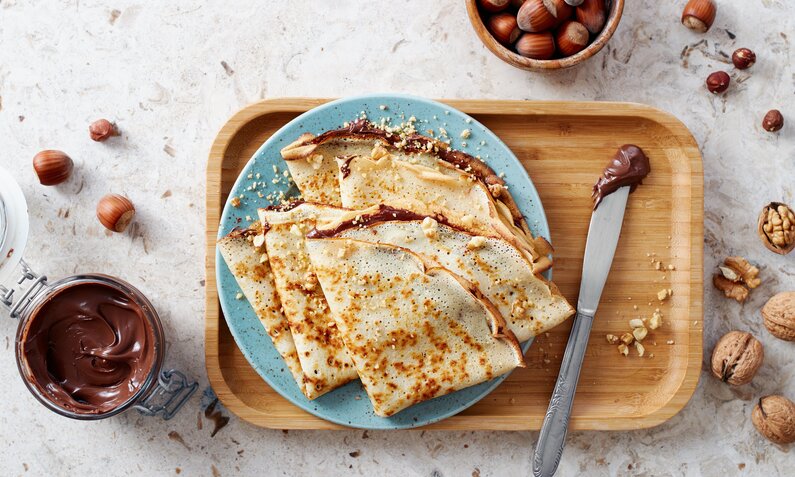 Zwei mit Nutella gefüllte Pfannkuchen auf einem blauen Teller angerichtet, daneben Nutella im Glas und viele frische Haselnüsse. | © Adobe stock / Melica