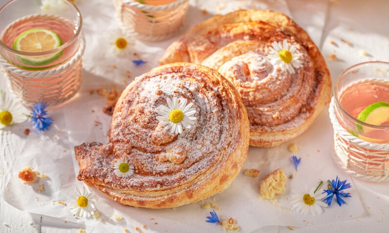 Zwei mit Puderzucker bestreute Hefeschnecken in sommerlicher Umgebung, daneben Limonaden in Gläsern. | © Adobe Stock / shaiith