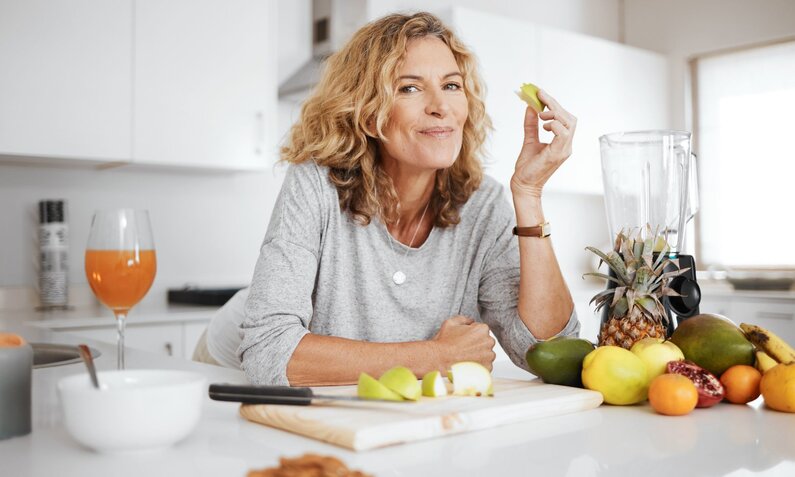 Eine lächelnde Frau mit lockigem, blondem Haar isst ein Stück Apfel in einer modernen Küche voller frischem Obst. | © Adobe Stock/ Nicholas Felix/peopleimages.com