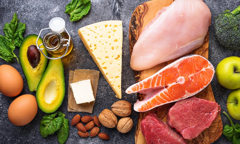 Frische Lebensmittel wie Avocado, Käse, Eier, Fleisch und Gemüse auf einer dunklen Granitplatte arrangiert. | © Adobe Stock/ Yulia Furman