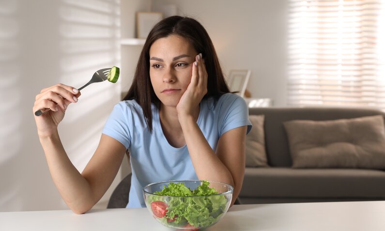 Junge Frau in hellblauem T-Shirt betrachtet gelangweilt eine Gurkenscheibe auf einer Gabel vor einer Schüssel Salat im hellen Wohnzimmer. | © Adobe Stock/ New Africa