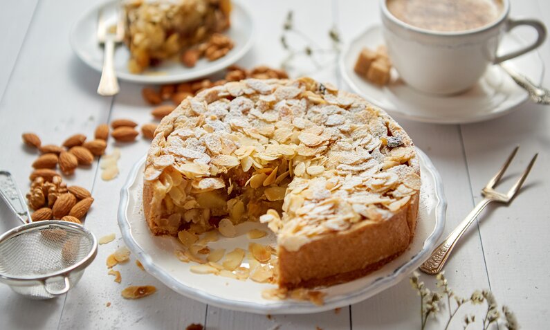 Angeschnittener Mandel-Joghurtkuchen auf einem weißen Teller, daneben eine Tasse Cappuccino und frische Mandeln. | © Adobe Stock / Dash