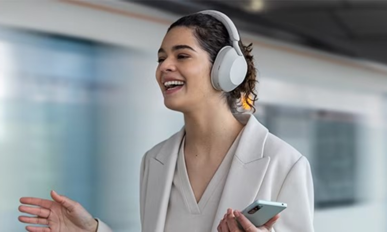 Fröhliche und entspannte Frau mit den Sony WH-1000XM5 Over Ear-Kopfhörer auf den Ohren. Der Hintergrund wird verschwommen dargestellt. | © Amazon/Sony