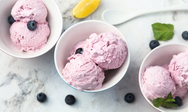 Drei Schalen mit rosa Heidelbeer-Eis, garniert mit Blaubeeren und Minzblättern, auf weißem Marmortisch. | © Getty Images/Mariha-kitchen