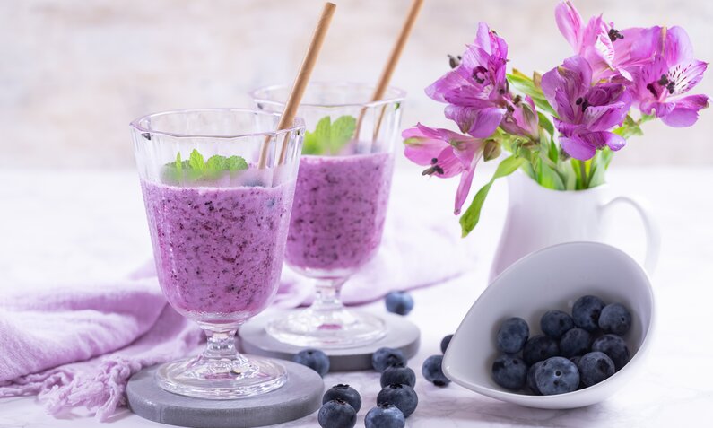 Zwei Gläser mit lila Blaubeersmoothie, garniert mit Minzblättern, neben einem weißen Krug mit violetten Blumen und einer Schale Blaubeeren. | © Getty Images/yumehana