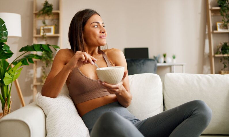 Eine junge Frau in Sportkleidung sitzt lächelnd auf einem Sofa und isst Müsli in einer gemütlichen, hellen Wohnung. | © Adobe Stock/ djile