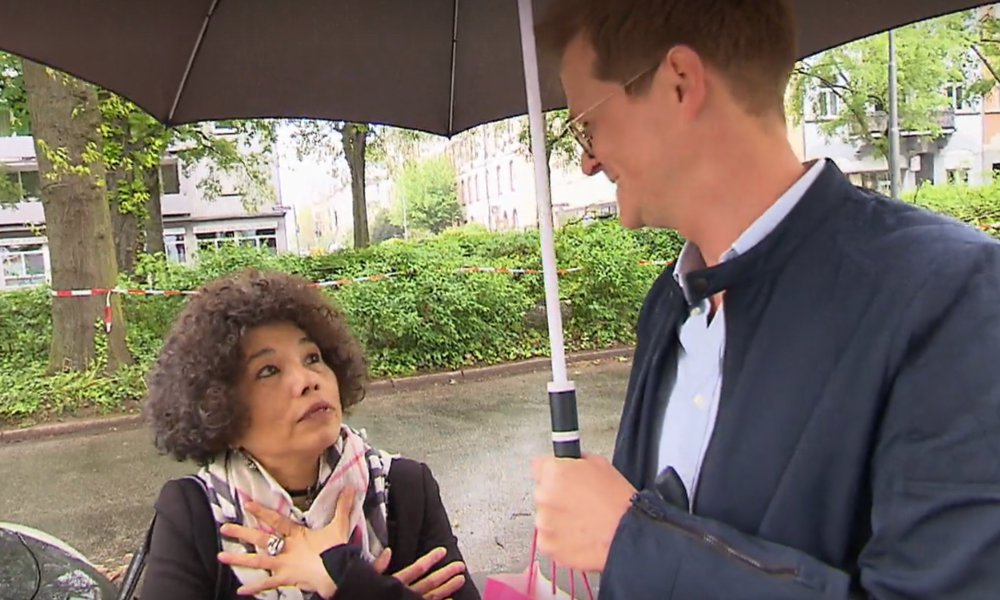 Ein Mann und eine Frau stehen unter einem Regenschirm, sie spricht gestikulierend, im Hintergrund ein parkähnliches Stadtgebiet. | © Screenshot Vox/RTL+ 