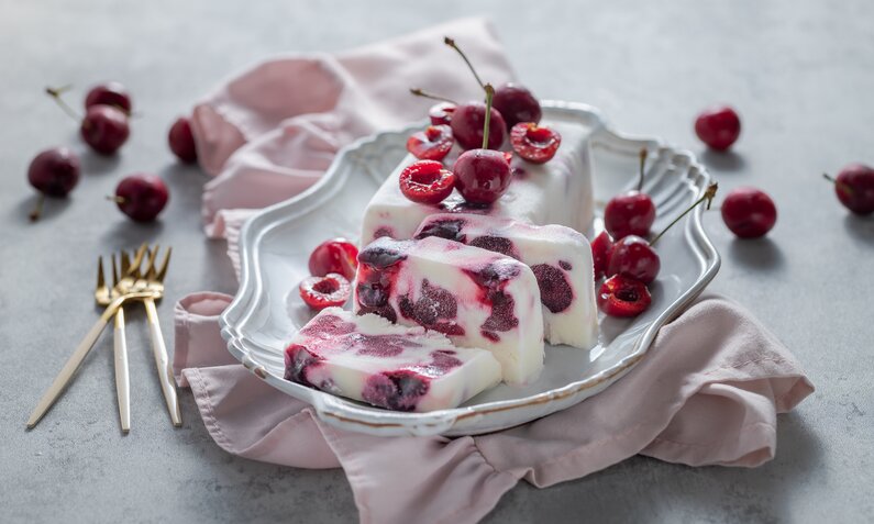 Angeschnittenes Kirsch-Joghurt-Parfait ohne Zucker mit frischen Kirschen auf einem weißen Teller angerichtet. | © Getty Images / 	yumehana