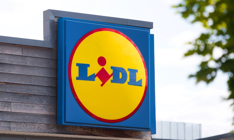 Logo der Supermarktkette Lidl in blau und gelb an einer Holzfassade bei Sonnenschein. | © Adobe Stock / Tobias Arhelger