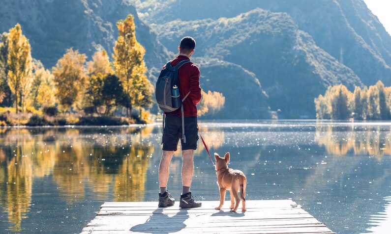 Mann in rotem Pullover und Rucksack steht mit Hund an der Leine auf einem Steg vor einem Bergsee. | © Adobe Stock / nenetus