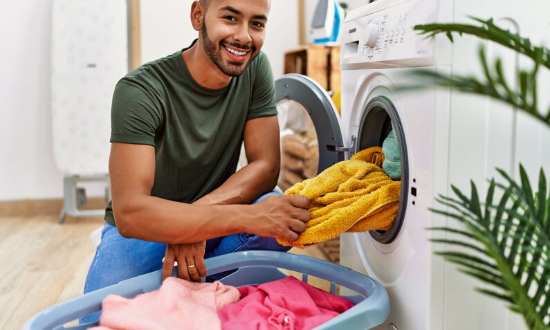 Ein junger Mann mit Kurzhaarschnitt und Bart legt lächelnd ein gelbes Handtuch in eine geöffnete Waschmaschine. | © Adobe Stock/ Krakenimages.com