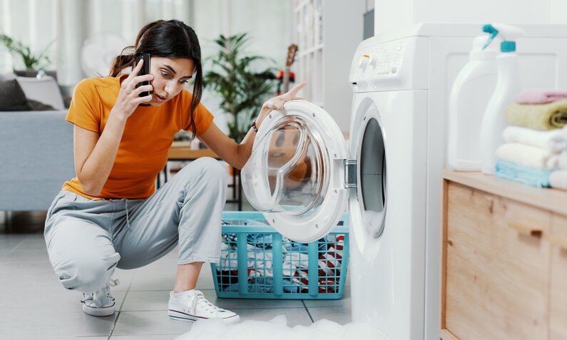 Frau mit dunklen Haaren in Sneakern telefoniert gestresst neben einer überlaufenden Waschmaschine im modernen Haushalt. | © Adobe Stock/ StockPhotoPro