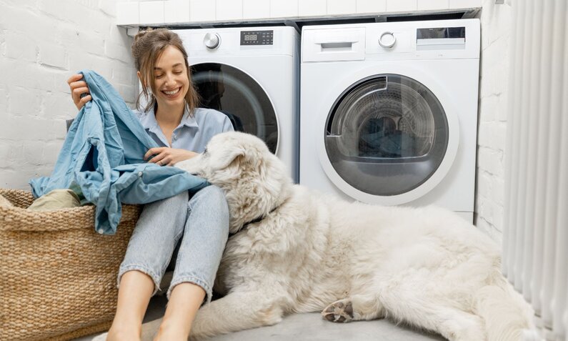 Junge Frau sitzt lächelnd auf dem Boden vor Waschmaschine, hält ein blaues Hemd und wird von großem, weißen Hund liebevoll angestupst. | © Adobe Stock/ rh2010