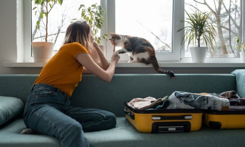 Junge Frau im orangefarbenen T-Shirt streichelt eine Katze auf der Fensterbank, während sie auf einem Sofa neben einem offenen Koffer sitzt. | © Adobe Stock / Maria