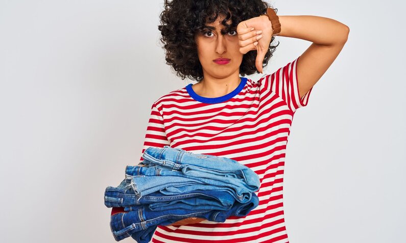 Junge Frau mit lockigem Haar zeigt einen Daumen nach unten, während sie missmutig einen Stapel Jeans in der Hand hält. | © Adobe Stock/ Krakenimages.com