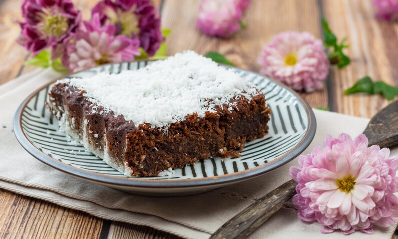 Ein Stück Kokos-Brownie auf einem gestreiften Teller, daneben frische lila Blumen und ein Holzlöffel. | © Adobe Stock / virginie