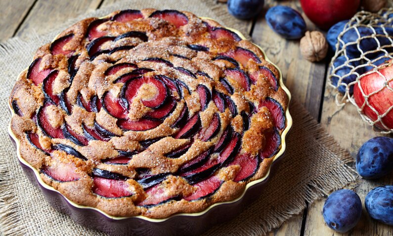 Low Carb Pflaumenkuchen in einer Kuchenform auf einem Holzuntergrund, daneben frische Pflaumen. | © Getty Images / GreenArtPhotography