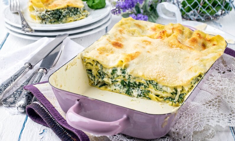 Low Carb Spinat-Lasagne in einer lila-Auflaufform, angeschnitten und dahinter auf einem Teller ein weiteres Stück Lasagne. | © Adobe Stock / HLPhoto