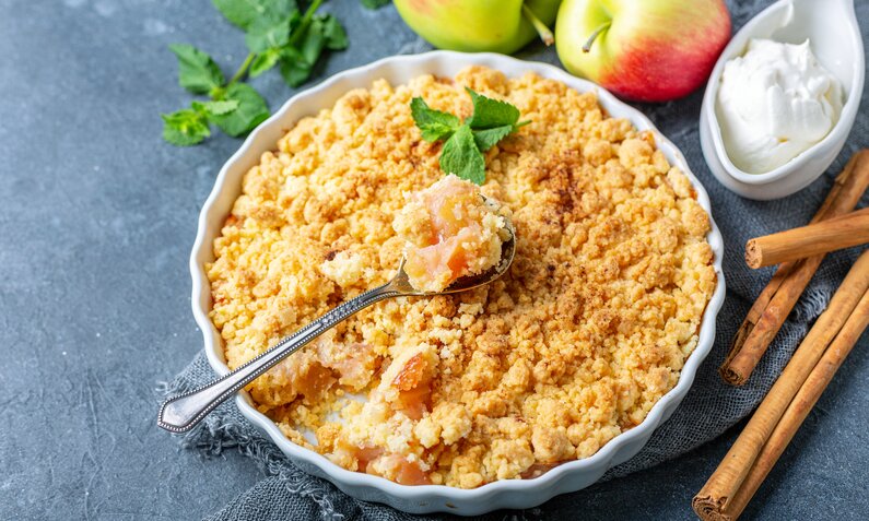 Apple Crumble in Auflaufform, mit einem Löffel drinnen stecken, daneben frische Äpfel, Eis und frische Minze. | © Adobe Stock / sriba3