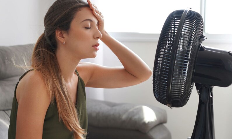 Junge Frau mit geschlossen Augen und Hand auf der Stirn sitzt erschöpft vor einem Ventilator im Wohnzimmer. | © Adobe Stock/ zigres