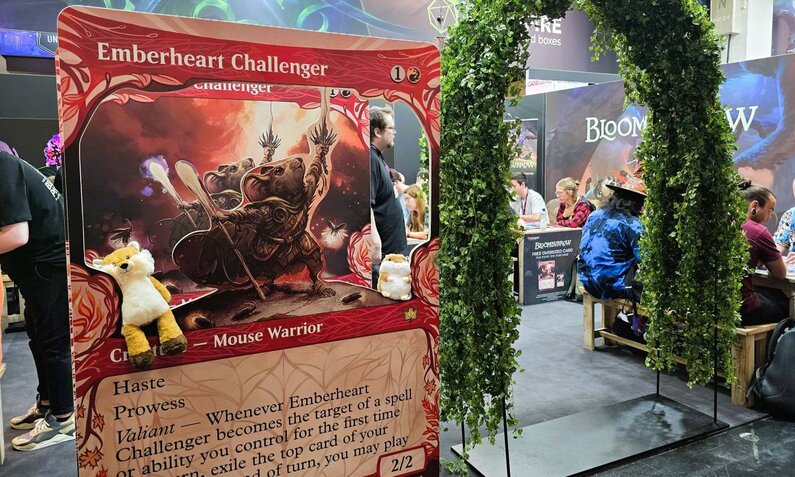 Messestand mit riesiger „Emberheart Challenger“-Karte und grünem Torbogen, dahinter Menschen, die konzentriert an Tischen sitzen und spielen. | © Sabrina Forst