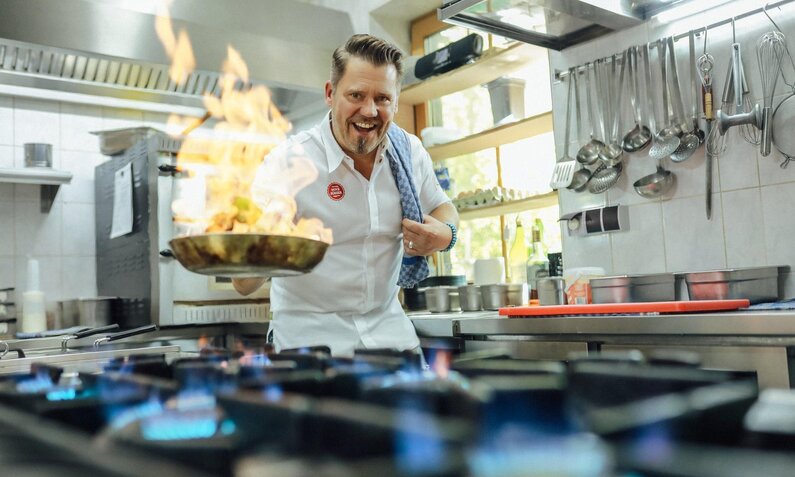 Mike Süsser trägt eine weiße Uniform und schwenkt eine brennende Pfanne über einem Gasherd in einer modernen Küche. | © Kabel Eins