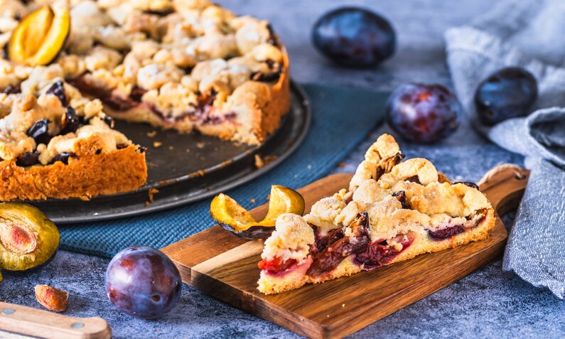 Angeschnittener Pflaumenkuchen auf einem Kuchenteller, im Vordergrund ein Stück des Kuchens auf einem weiteren Teller. | © Getty Images / Daniela Baumann