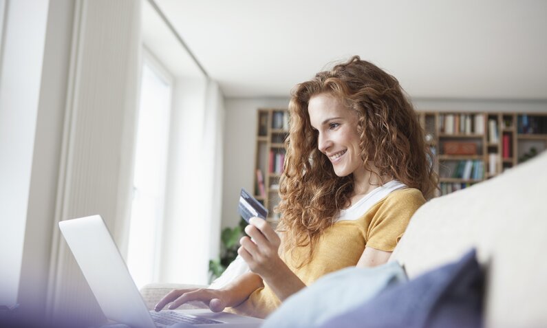 Junge Frau mit lockigem, rotbraunem Haar sitzt lächelnd auf dem Sofa und hält eine Kreditkarte vor dem Laptop in einem hellen Wohnzimmer. | © Getty Images/Westend61